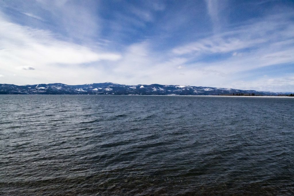 Photographs of the lake at Wayfarer's Park  where the water in the lake ripples towards the bottom right corner. There are dark blue mountains along the whole picture in the background with thin white clouds gently streaming across the blue sky.