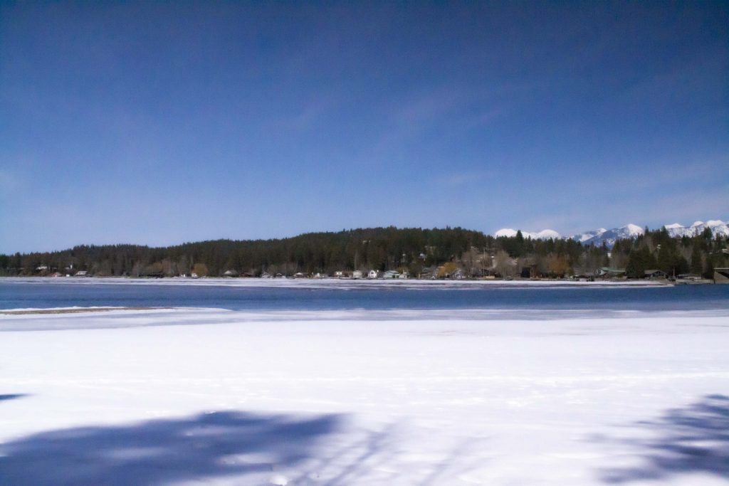 Photo of snowy foreground with a little strip of Flathead Lake in the center and lots of trees and small houses in the background. There is a deep blue sky with very few clouds.