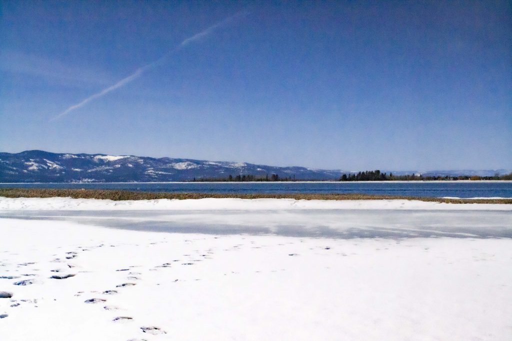 Photo of the lake at Wayfarer's Park with a lot of snow and ice on the foreground with footprints pressing into the snow. There is a jet stream in the sky and some mountains in the background.