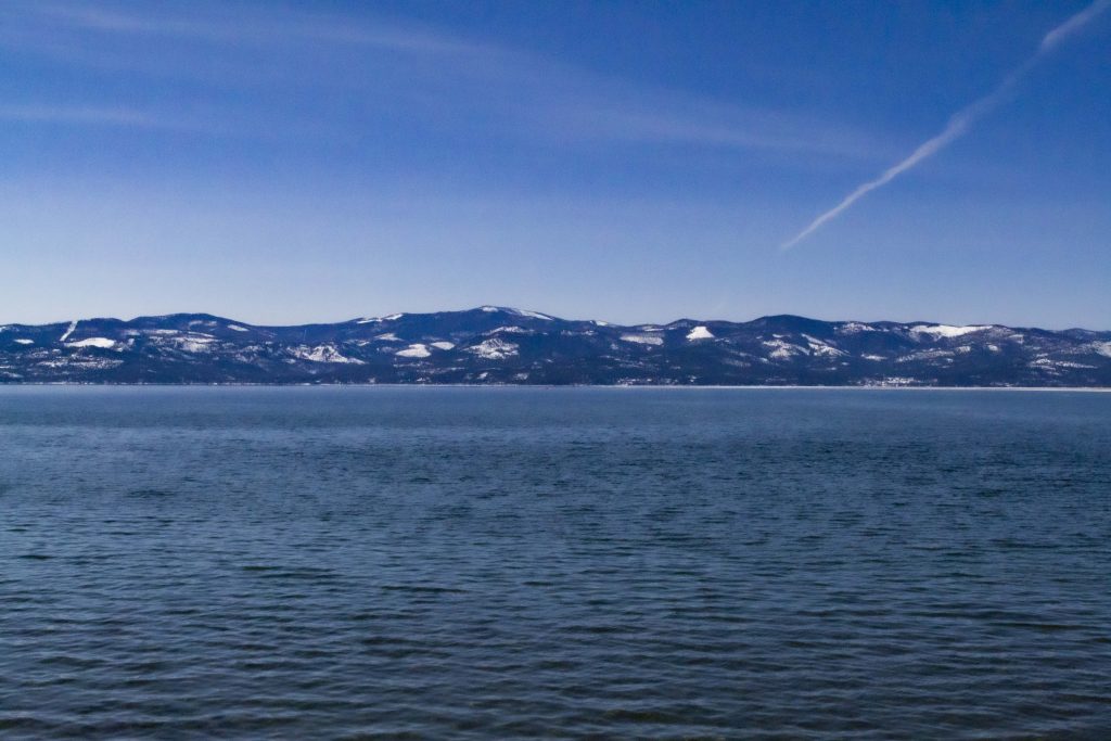 Photographs of the lake at Wayfarer's Park of deep blue water that is rippling. There are dark blue mountains way off in the background with a crisp blue sky and one lonesome jet stream in the upper right corner.