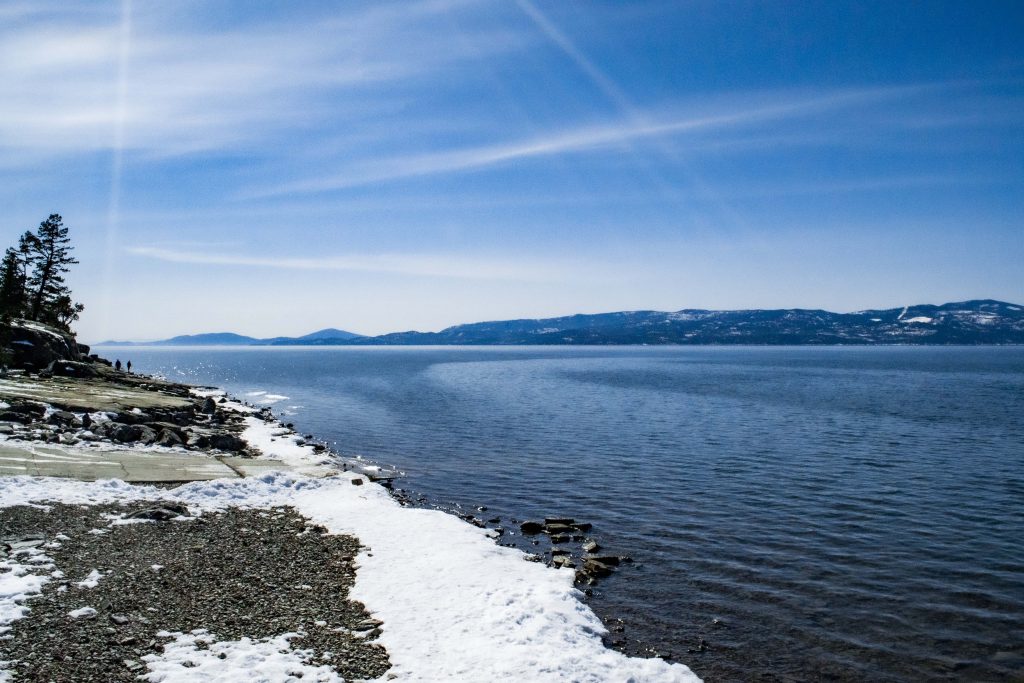 Image of the boat dock on the left and a piece of Flathead Lake on the right two-thirds of the picture. The water and sky are a deep blue and some sunshine rays shine in the sky. There are two people walking along the water's edge in the very back of the background.