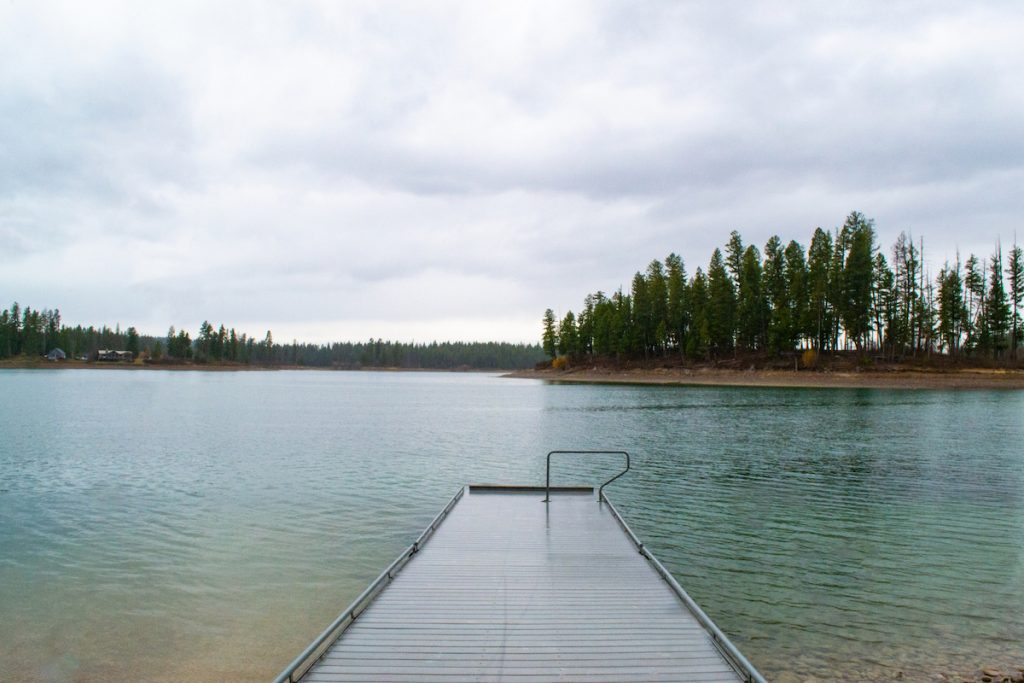 Photo of Echo Lake in the Flathead Valley where a gray dock is in the center bottom that leads the eye into the lake. The lake is a mixture of green and blue where the rocks are visible nearest the viewer. There are small islands in the background with sandy beaches and trees in the background. There is one such island in the middle right of the photo that creates an inlet of the lake. The sky is filled with puffy, purplish gray clouds that loom over the scene.