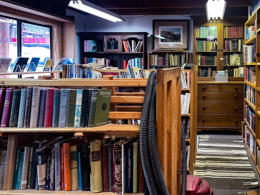 Left side of store that is through a doorway after you walk in. There are many bookcases and shelves of books. There are a couple isles of books and a vacuum is hanging over one of them. There are also books displayed in the store's window on the leftmost side.