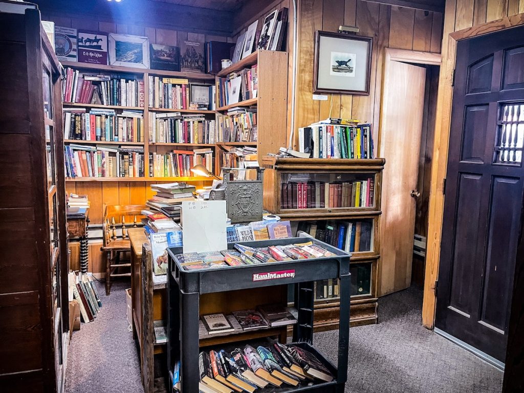 The right side of the store after you come in with a table, a cart, a couple of bookcases, and a wall displaying lots of books. There is a brown, wooden theme for all of the pieces