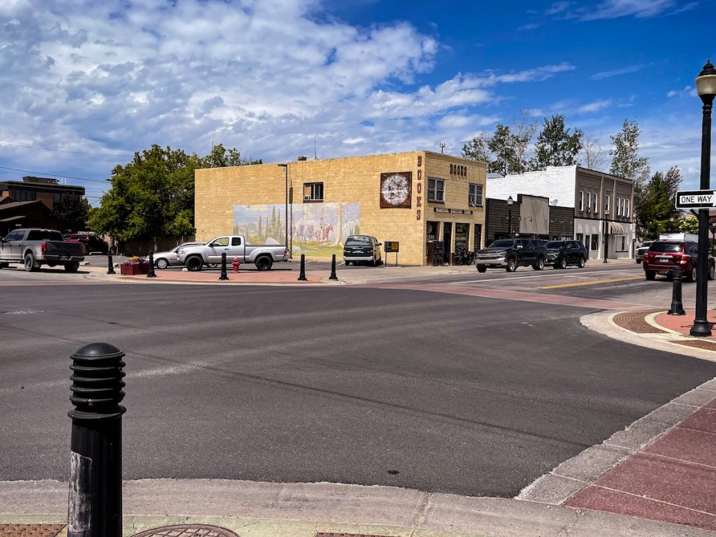 A picture of Blacktail Mountain Books - a used bookstore in Kalispell. There is a mural of a cowboy riding a horse among some trees on the side of a brown building that has "BOOKS" along the side of it. The picture is taken from the outside of a street intersection.