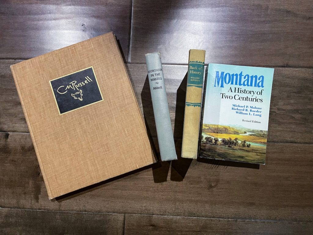 four of the five books I got from Bay Books and Prints bookstore in Bigfork Montana. They are all placed on wooden floor. The left most books is a large tan book about C M Russell and the right most is a newer book on Montana History while there are two older books placed so that their spines face the camera.