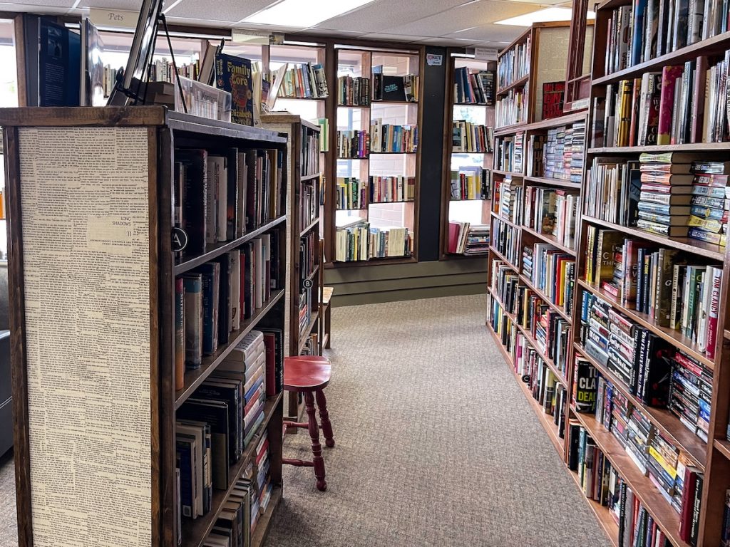 Looking down an isle of books with a taller bookcase on the right and a shorter one on the left with a long bookcase of books that reaches the ceiling in the back. This is next to the reading area on the picture on the left.