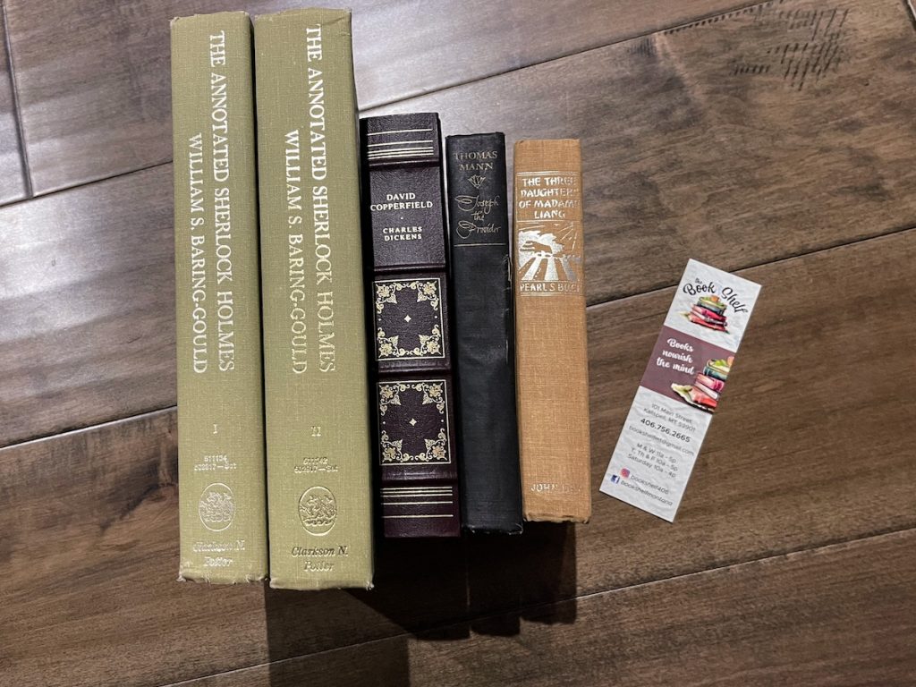 A selection of two Sherlock Holmes books, an old-fashion binding of "David Copperfield" in purple, "Joseph the Provider" in black and "The Three Daughters of Madame Lang" in orange sitting on wooden floor next to a bookmark from The Bookshelf.