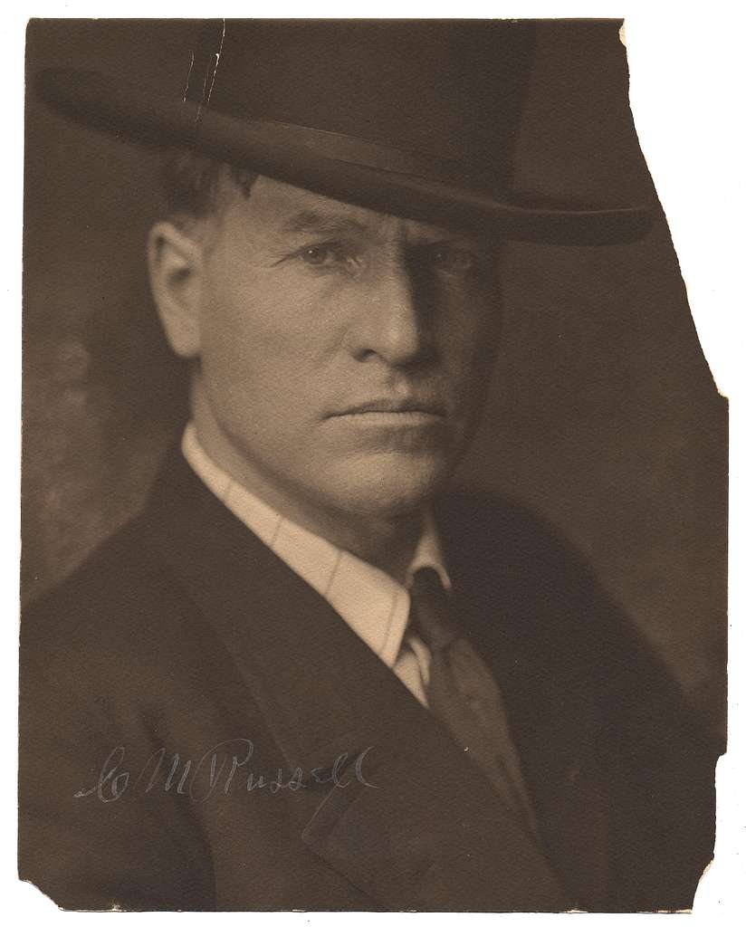 A portrait of C M Russell. It has a brown sepia look to it. Russell is looking at the photographer while wearing a dressy cowboy hat and in a nice suit.