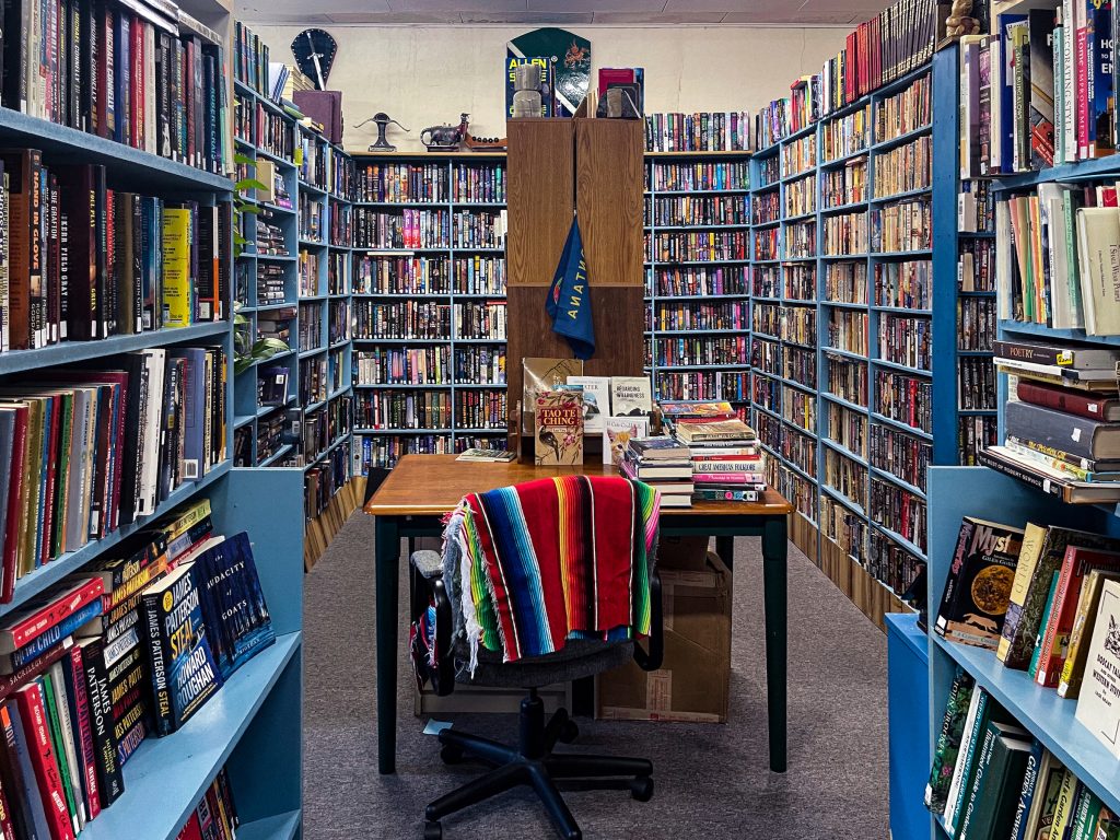 A table stacked full of books in between isles of bookshelves full of books